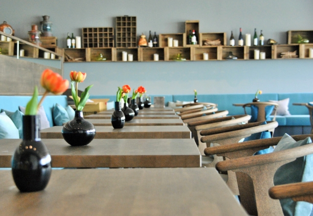 Frühstücksrestaurant Leev im SeeHuus. Moderne Möbel aus Holz. Sitzbank türkis gepolstert. Kissen in türkis und blau. Regal mit maritimer Dekoration und Weinflaschen an der Wand. Tulpen in schwarzen Vasen auf den Tischen.